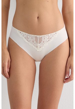 Femme portant un sous-vêtement style bikini blanc orné de dentelle, debout devant un fond clair uni, montrant le ventre et le haut des cuisses.
