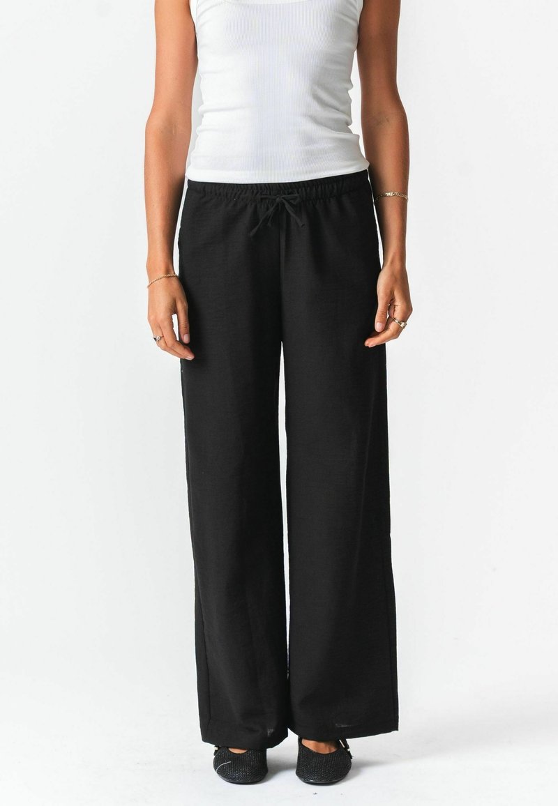 Femme portant un pantalon large noir avec une taille à cordon, un haut blanc sans manches et des chaussures noires sans lacets, debout devant un fond blanc.