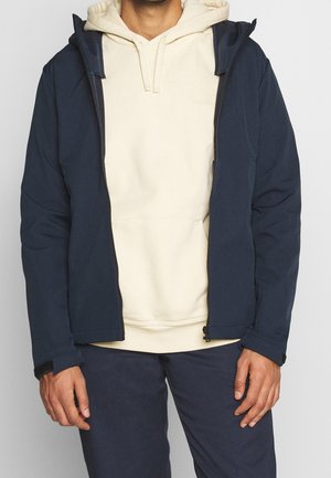 Mann trägt beige Kapuzenpullover unter einer aufgezogenen marineblauen Jacke mit passender navyblauer Hose, steht vor einfachem Hintergrund.