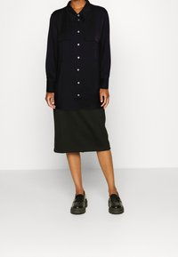 Camicia lunga blu navy con chiusura a bottoni, due tasche sul petto e gonna nera fino al ginocchio abbinata a mocassini neri lucidi. Tessuto morbido.