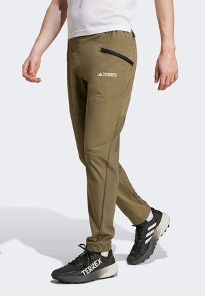 Homme portant un pantalon Terrex vert olive avec une poche zippée et des baskets Terrex noires avec des bandes blanches, debout sur un fond blanc.