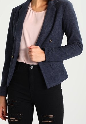 Kobieta ubrana w marynarkę koloru granatowego na lekkiej różowej bluzce i czarnych, podartych dżinsach, trzymająca jedną ręką klapę marynarki.