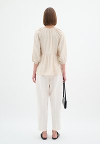 Top peplum beige con maniche a sbuffo, abbinato a pantaloni crema. La modella tiene una piccola borsa nera e indossa scarpe nere basse.
