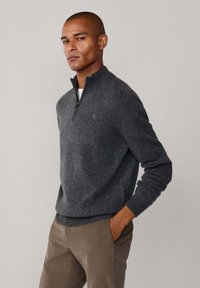 Pull en laine gris à col zippé 1/4 avec une finition texturée, poignets et ourlet côtelés, porté sur une chemise blanche, associé à un chino beige.