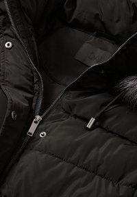 Svart vadderad jacka inuti med en silverfärgad dragkedja och tryckknappar, med en mjuk pälsfodrad huva och tygmärke inuti.