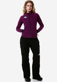 Giacca in fleece di colore viola con colletto alto e zip frontale, abbinata a pantaloni neri con tasche zippate e dettagli con logo.