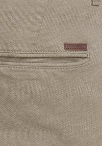 Tissu beige texturé avec une petite poche horizontale et une étiquette en cuir marron embossée du texte « JACK & JONES ».