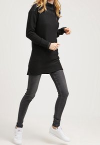 Un maglione tunica nero a maniche lunghe abbinato a jeans skinny grigi scuri e sneakers bianche. Il maglione presenta un collo alto e spacco laterale.