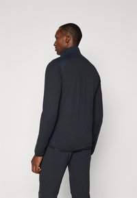 Un homme de profil porte une veste zippée sombre avec des manches texturées et un pantalon sombre assorti, sur un fond clair et uni.
