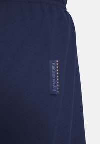 Marineblaue Baumwoll-Jogginghose mit elastischem Bund, ausgestattet mit einem seitlichen Etikett in Blau mit roten und weißen Streifen. Glatte Textur, minimalistisches Design.