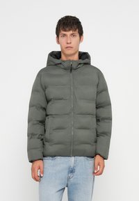 Παλτό puffer σε ελαιόλαδο πράσινο, με κλείσιμο φερμουάρ μπροστά, κουκούλα και τετράγωνη γραμμή. Κατασκευασμένο από μαλακό, ραμμένο υλικό με δύο μπροστινές τσέπες.