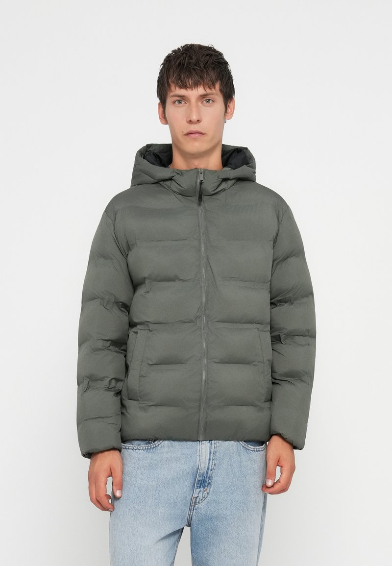 Παλτό puffer σε ελαιόλαδο πράσινο, με κλείσιμο φερμουάρ μπροστά, κουκούλα και τετράγωνη γραμμή. Κατασκευασμένο από μαλακό, ραμμένο υλικό με δύο μπροστινές τσέπες.