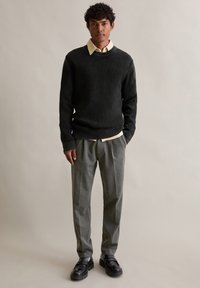 Tmavě zelený pletený svetr s kulatým výstřihem, přes který je navlečený světlý béžový košilový top, doplněný šedými zakázkovými kalhotami a černými botami.