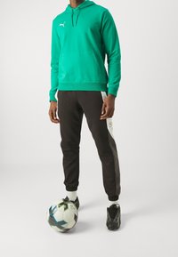 Felpa verde con tasca frontale e logo bianco, pantaloni jogger neri con pannello laterale bianco e scarpe da calcio nere accanto a un pallone da calcio.