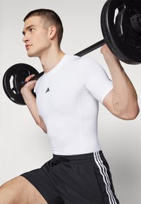 Hvid tætsiddende atletisk skjorte med sort logo, parret med sorte shorts med tre hvide striber. Model løfter en vægtstang.