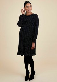 Vestido de maternidad negro, con textura, de mangas largas, cinturilla y largo hasta la rodilla. Llevado con medias negras y botas de tacón puntiagudas hasta el tobillo.