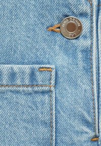 Svetlomodrá denimová tkanina s niťou šedej farby označenou ako "KING DENIM" a kontrastným béžovým prešívaním, s obdĺžnikovou kapsou a rozstrapkaným detailom.