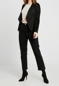 Veste en suédine noire avec un design à ouverture frontale, associée à un haut beige clair et un jean noir. Le modèle porte des bottines à talons aiguilles noires.