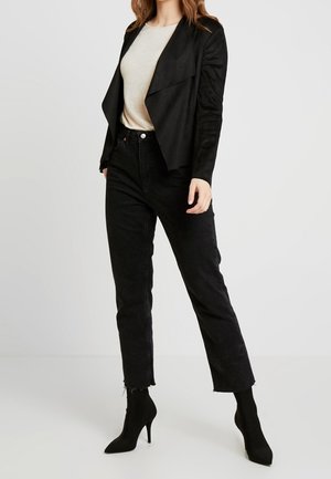 Chaqueta negra de ante sintético con diseño de frente abierto, combinada con una camiseta beige claro y vaqueros negros. La modelo lleva botas de tacón aguja negras.