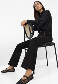 Schwarze Bluse mit langen Puffärmeln, hochgeschnittene schwarze Schlagjeans und schwarze Mesh-Ballerinas, die auf einem schwarzen Stuhl mit geflochtenem Sitz sitzt.