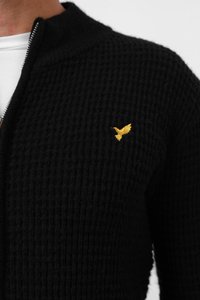 Cardigan noir en tricot texturé avec un col montant et une fermeture éclair argentée. Présente un petit logo d'oiseau brodé doré sur la poitrine.