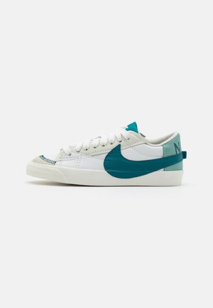Niskie białe sneakersy z turkusowym logo Nike, turkusową zakładką na pięcie, białymi sznurówkami i kremową podeszwą na jednolitym tle.