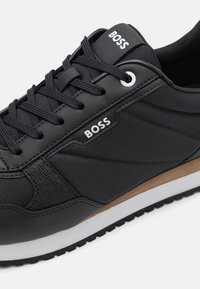 Zapatilla negra de material sintético liso, con paneles laterales texturizados, detalles en blanco y una suela exterior marrón a contraste.