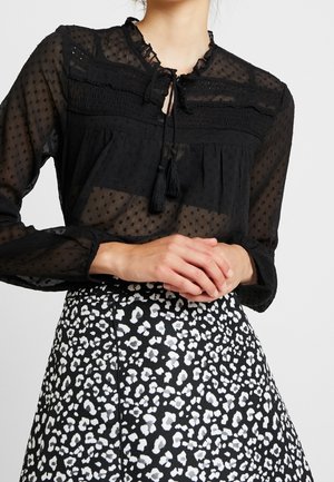 Zwarte doorschijnende blouse met lange mouwen, een gestippelde textuur en kwastjeskoord, gecombineerd met een zwart-witte geruite hoge taille rok.
