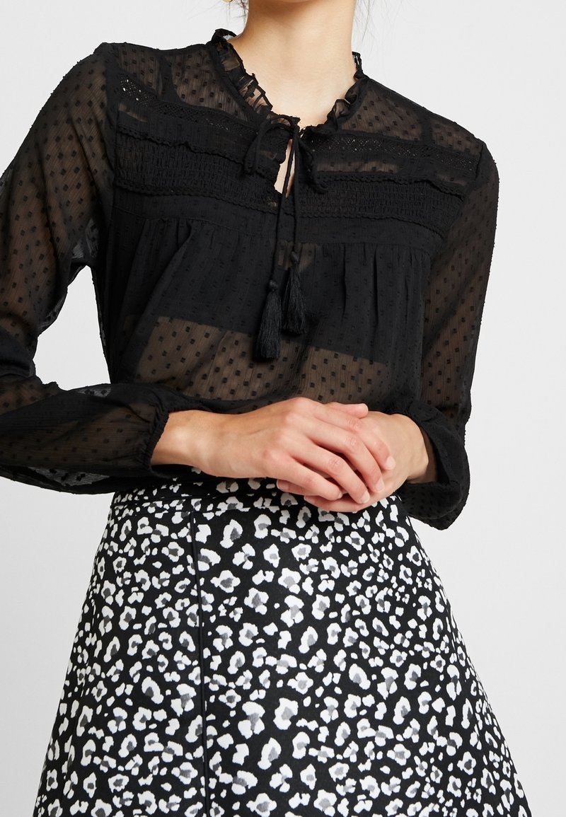 Black sheer long-sleeve blouse with dotted texture and tassel ties paired with a high-waisted black and white spotted skirt.