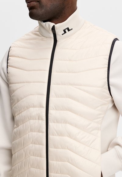 Uomo che indossa un gilet trapuntato beige chiaro con cerniera nera e logo sul colletto alto, sopra una camicia bianca a maniche lunghe.