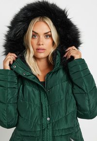 Grön pufferjacka med kapuschong av svart konstpäls. Har en vadderad textur, dragkedja och snap-knappar, samt sidofickor.