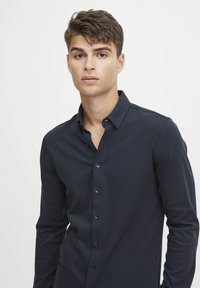 Cămașă bleumarin cu mâneci lungi, fabricată dintr-un material neted. Dispune de un guler clasic și nasturi negri, cu o croială ajustată.