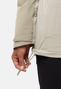 Beige buitenjas met een gladde textuur; hand die de koord aan de onderkant verstel; heeft ritszakken en een getailleerd ontwerp.