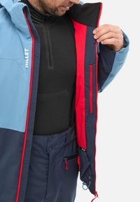 Blue and navy ski jacket with red accents, zippered pockets, and textured inner lining. Features prominent brand logo "MILLET" on the exterior.