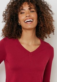 Röd v-ringad tröja med långa ärmar, gjord av ett mjukt material. Har ribbade detaljer runt halsringningen. Modellen visar ett glatt uttryck.