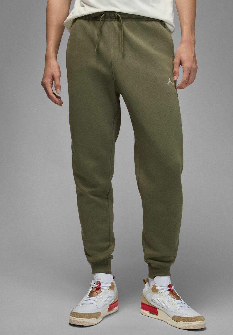 Persona che indossa pantaloni jogger verde oliva con vita con coulisse e sneaker alte bianche, beige e rosse su sfondo grigio.