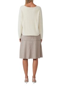 Vrouw gezien van achteren, gekleed in een donzige witte lange mouwen trui, een beige doorgelofte suède rok tot op de knie en witte hakken.