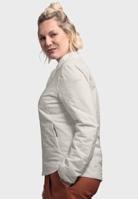 Light gray quilted jacket with a stand-up collar and zippered front. Features side pockets and a logo on the left sleeve. Satin texture.