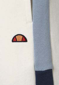 Jersey de algodón con un cuerpo color crema y acentos en azul claro y azul marino. Destacado logo bordado en naranja, rojo y amarillo en la parte izquierda.