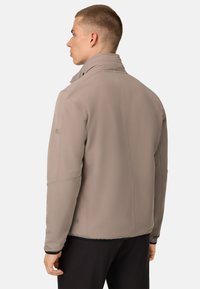Veste softshell beige avec un col haut, fermeture éclair et poignets. Présente un détail de logo subtil sur la manches et une surface texturée.