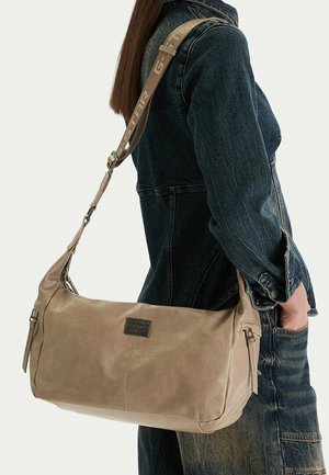 Osoba v tmavé džínové bundě držící béžovou koženou kabelku přes rameno s nastavitelným popruhem a viditelným logem značky.