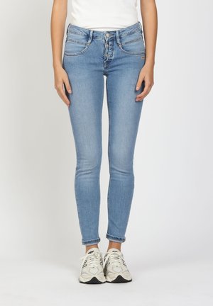 Slim-fit light blue jeans with button fly worn by a person in white sneakers and a white shirt against a plain background.