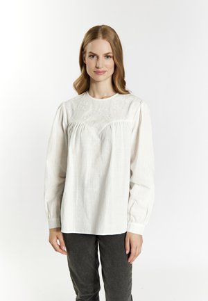 Donna con capelli castano chiaro che indossa una camicetta bianca a maniche lunghe con ricami e pantaloni grigio scuro, in piedi davanti a uno sfondo bianco uniforme.
