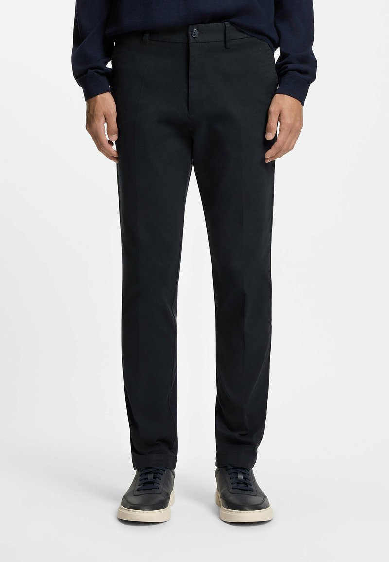 Homme portant un pantalon noir sur mesure, un pull bleu marine et des baskets noires à semelles blanches, debout devant un fond blanc uni.