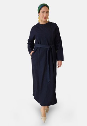 Mujer con un vestido largo azul marino con cinturón, pañuelo verde en la cabeza, grandes pendientes dorados y zapatos beige puntiagudos, de pie con una mano en el bolsillo.