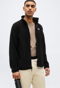 Νεαρός άνδρας που φοράει μαύρο φλις σακάκι με φερμουάρ, μπεζ πουκάμισο και ανοιχτόχρωμο παντελόνι, κρατώντας ένα μαύρο παγούρι με νερό μπροστά σε απλό φόντο.