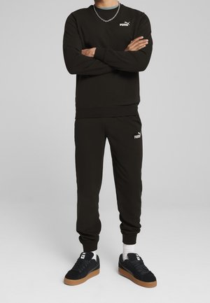 Homme portant un sweat-shirt Puma noir et un jogging, baskets noires avec semelles en gomme, debout les bras croisés devant un fond gris uni.