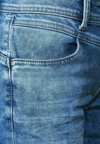 Světle modré džíny z denimu s viditelným prošíváním, dvěma zadními kapsami a malým kulatým kovovým detailem ve tvaru knoflíku. Textura vypadá měkce a opotřebovaně.