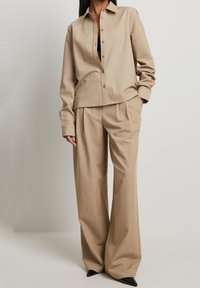 Chemise beige ajustée avec des boutons et un col, associée à un pantalon large plissé assorti. Texture lisse et silhouette nette. Chaussures noires.