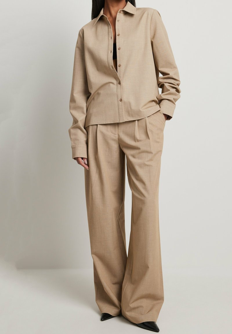 Chemise beige ajustée avec des boutons et un col, associée à un pantalon large plissé assorti. Texture lisse et silhouette nette. Chaussures noires.
